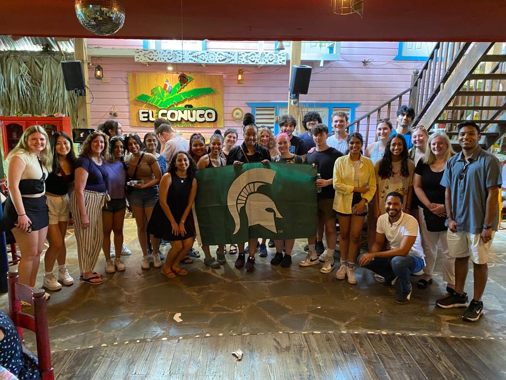A group of people at a restaurant with a disco ball holding up an MSU flag