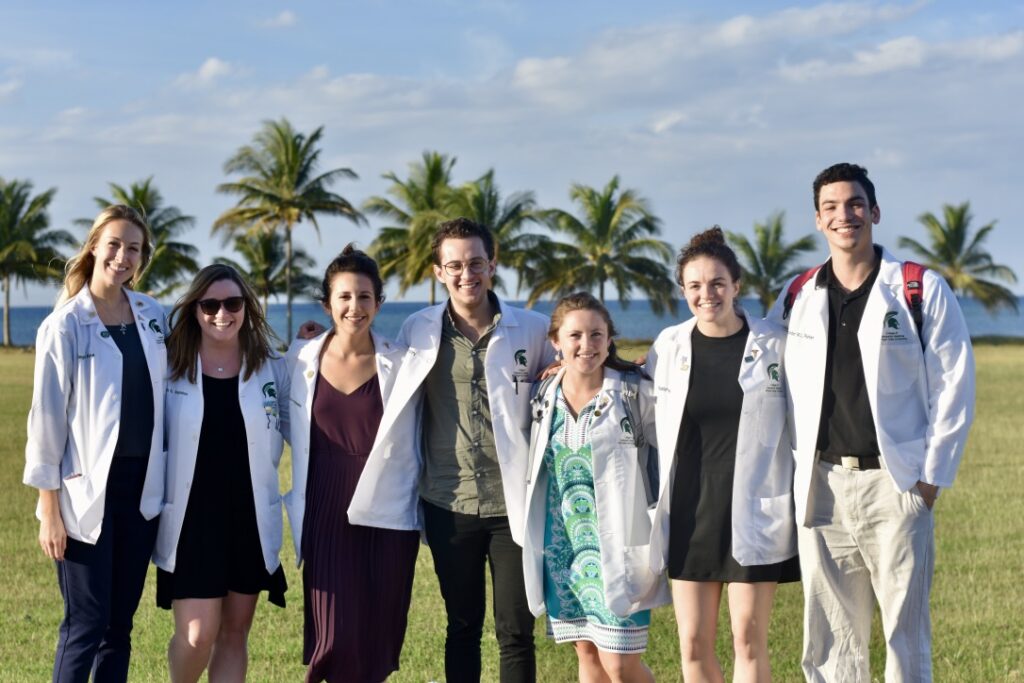A group of people in white lab coats smiling in front of palm trees with water in the distance