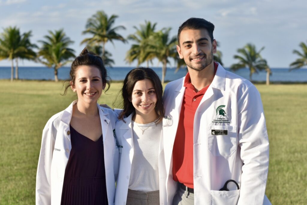 Three people in white lab coats smiling in front of some palm trees with water in the distance