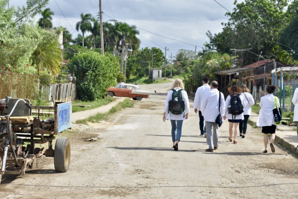 A group of people with backpacks walking down a road with trees and palm trees and various old structures along the street.