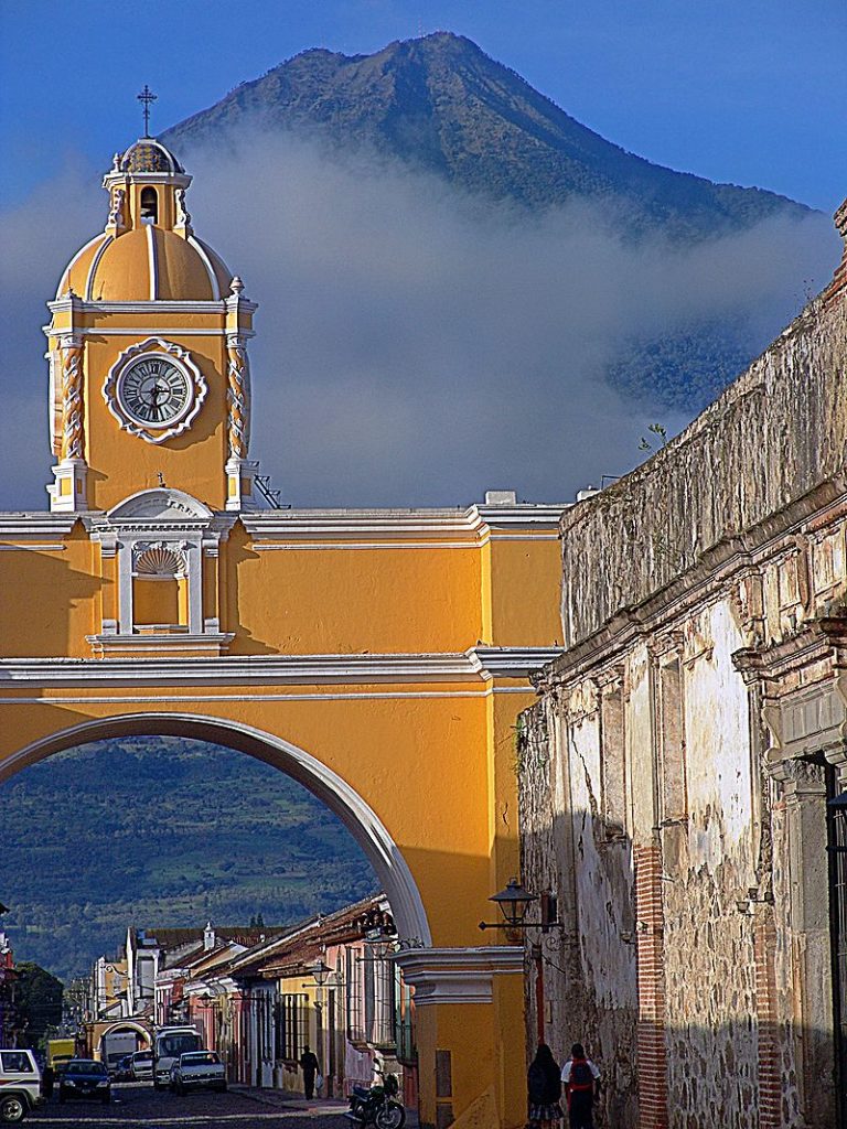 A yellow clock tower with a turret on top of an archway that is in front of a mountain.