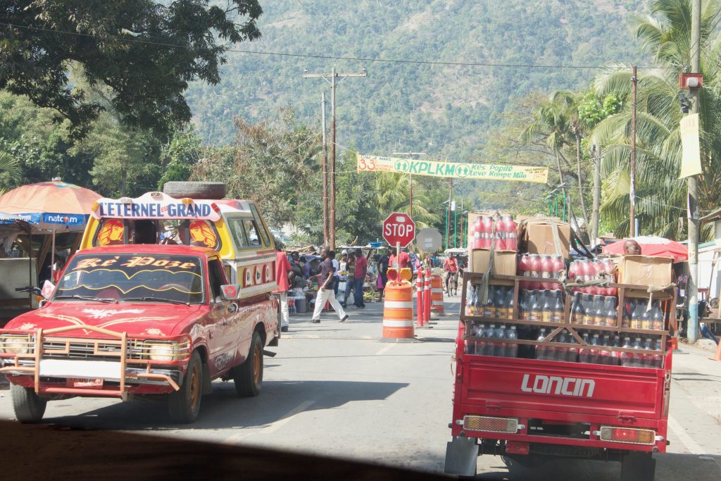 Two trucks, one with lots of bottles on it, both are red, are on a road with trees in the distance